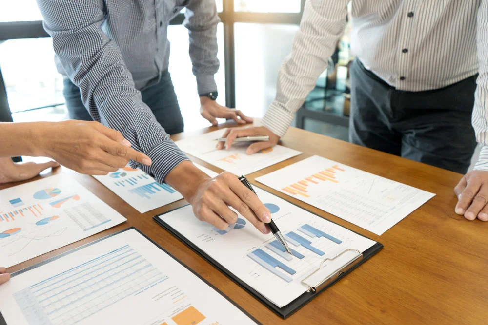 A group of three people standing over a table with sheets of paper showing bar graphs and pie charts, with one of them pointing at a bar graph with a pen