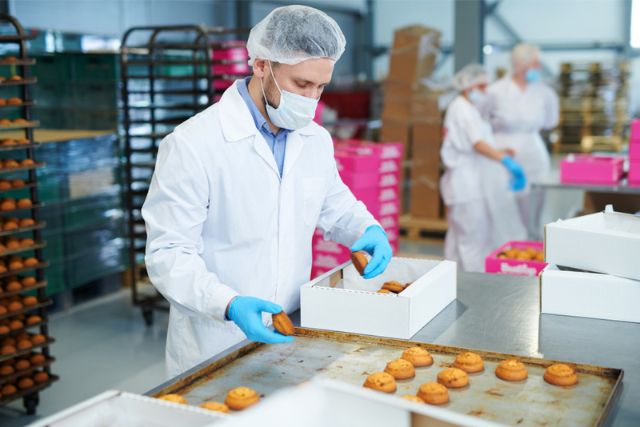 Confectionery factory worker in white coat collecting freshly baked pastry from tray and putting it into paper box