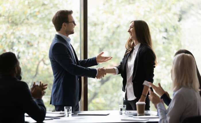 Business professionals shaking hands during a meeting, with colleagues seated and clapping around the table