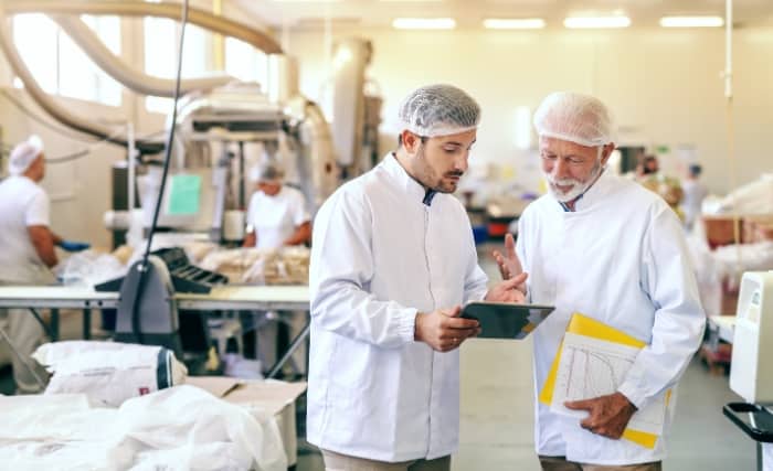 Two food factory workers in lab coats discussing information on a tablet