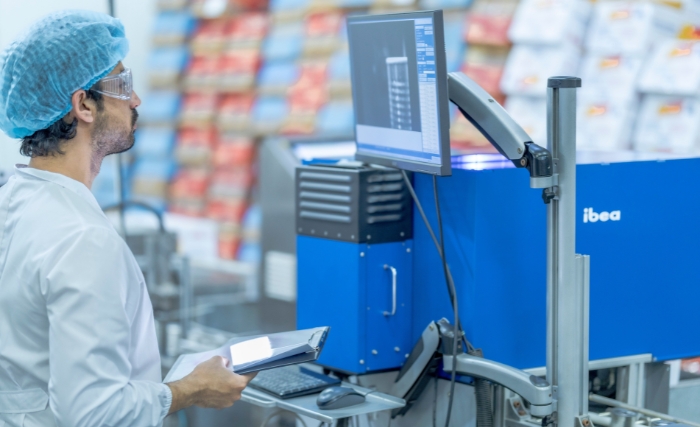 A worker in a clean white coat, hairnet, and protective glasses operates machinery in a food production facility. He’s holding a tablet while observing data on a computer monitor connected to blue industrial equipment, ensuring process quality and efficiency.