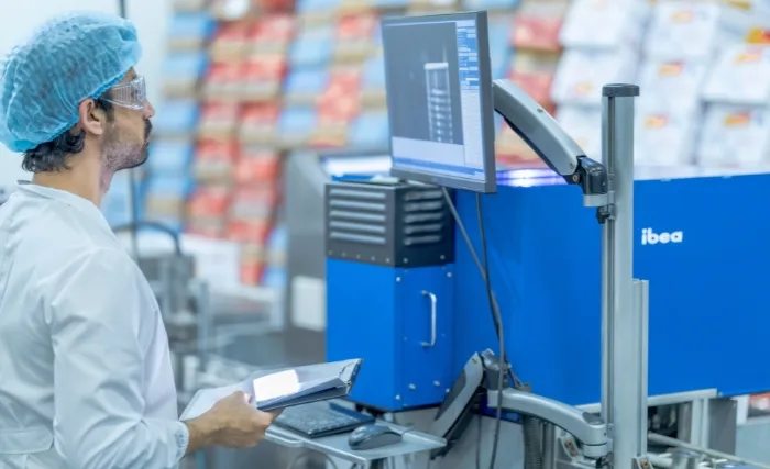 A worker in a clean white coat, hairnet, and protective glasses operates machinery in a food production facility. He’s holding a tablet while observing data on a computer monitor connected to blue industrial equipment, ensuring process quality and efficiency.