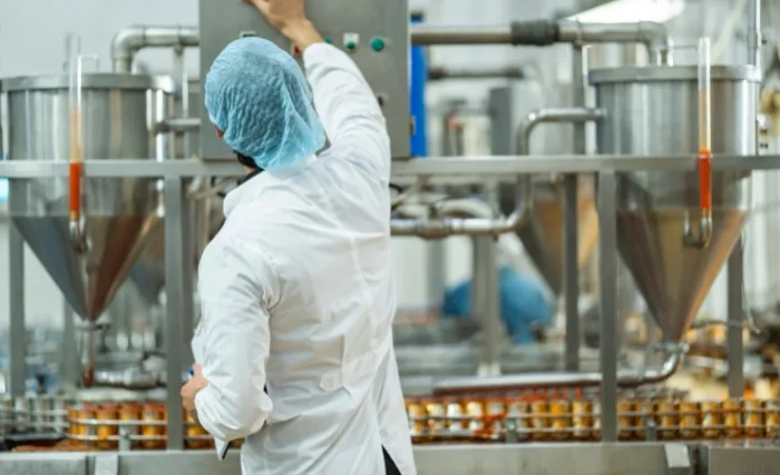 A worker wearing a white lab coat and blue hairnet operates stainless steel machinery in a modern food processing plant. He adjusts the control panel overseeing automated filling lines producing food items in jars or containers.