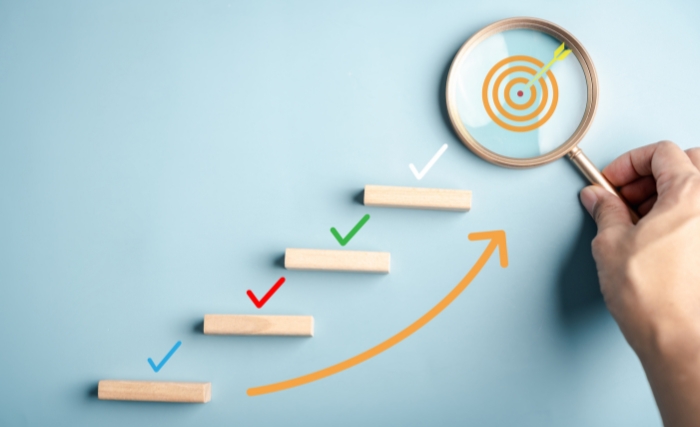 Measuring marketing performance: Visual of stepped progress blocks with ticks and arrows leading to a target under a magnifying glass