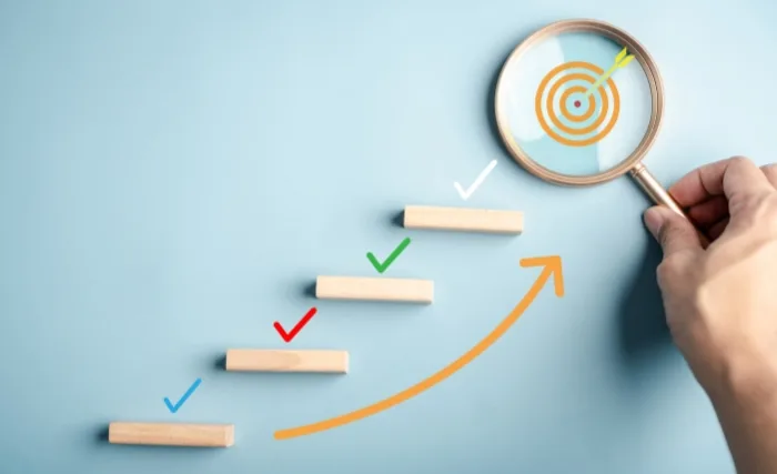 Measuring marketing performance: Visual of stepped progress blocks with ticks and arrows leading to a target under a magnifying glass
