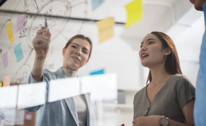Operational cost reduction, Two professionals discussing ideas in front of a glass board covered in notes and diagrams, illustrating operational cost reduction through collaborative planning.