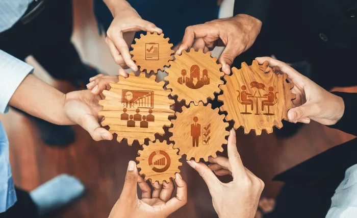 resource allocation for business growth: Multiple hands connecting wooden gears with business icons, symbolising collaboration, resource allocation, and coordinated business growth strategy.