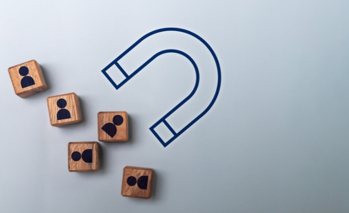 Improve lead quality: Five wooden blocks printed with people icons arranged next to a drawn blue horseshoe magnet on a light grey background, illustrating the concept of attracting customers.