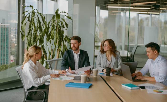 Using AI tools in B2B strategy and consulting UK Four business professionals sit around a meeting room table in a modern glass-walled office, reviewing printed charts and documents, with laptops and coffee cups on the table and a large plant in the background.
