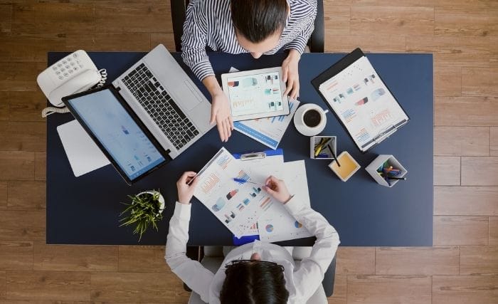 When should you hire an interim marketing director: An overhead view of two people at a dark desk reviewing printed charts and graphs alongside a laptop, tablet, coffee, and stationery.