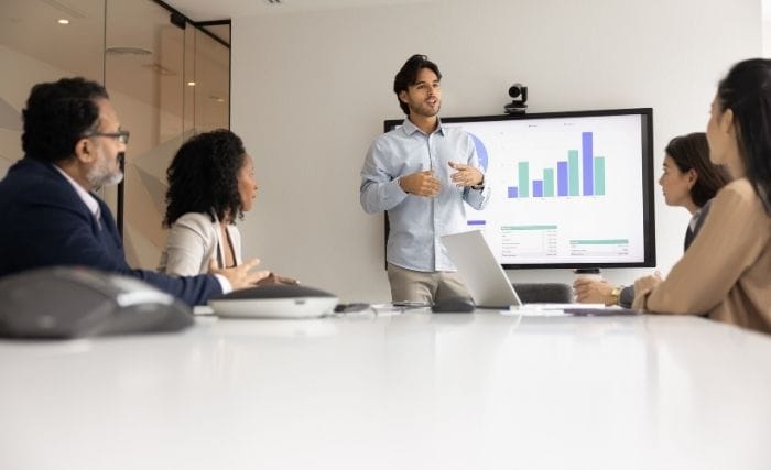 Interim commercial director: A man presenting bar chart data on a large screen to four colleagues seated around a boardroom table.