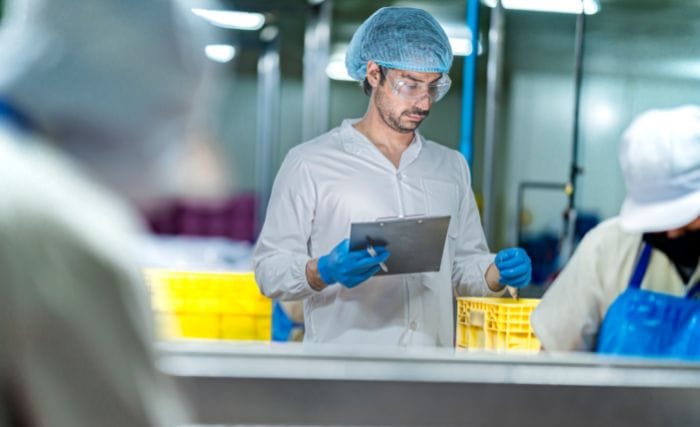 is AI worth it for food production: A man in a hairnet, safety glasses and blue gloves holding a clipboard while inspecting food on a production line, with a colleague in a hairnet visible in the foreground and yellow crates behind him.