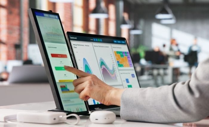 When should you hire an interim director: A person in a grey top pointing at a touchscreen monitor displaying a business analytics dashboard with charts and graphs, in a modern open-plan office.