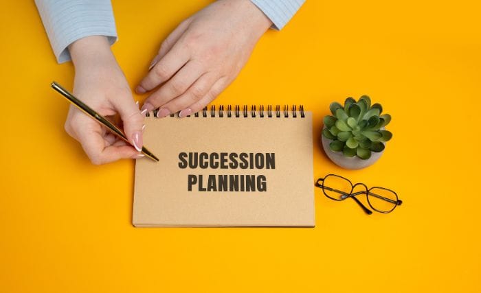 Succession Planning for Family-Owned Manufacturers: A person writes with a gold pen on a kraft-paper spiral notebook with “Succession Planning” printed on the cover. The notebook sits on a bright yellow desk beside a small succulent in a pot and a pair of glasses.