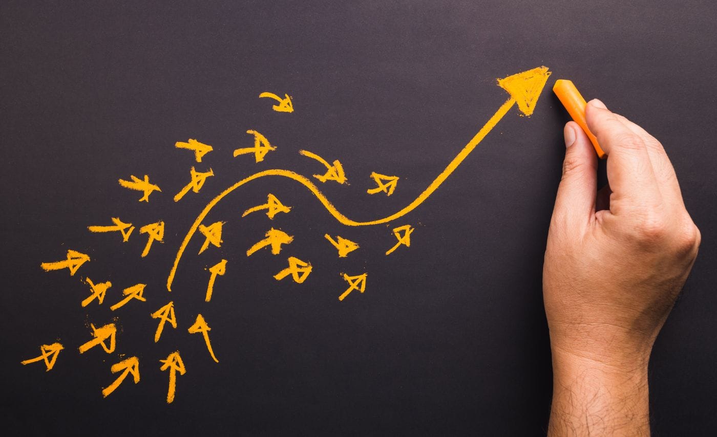 Why Are Your Marketing Leads Not Converting: Hand drawing an orange chalk arrow that curves upward out of a scattered cluster of small arrows pointing in different directions on a blackboard.