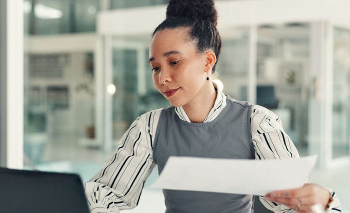 preparing a manufacturing business for the next generation: A young woman with her hair in a bun reads a sheet of paper at her desk in a bright office with glass walls. She’s wearing a grey pinafore over a striped blouse.